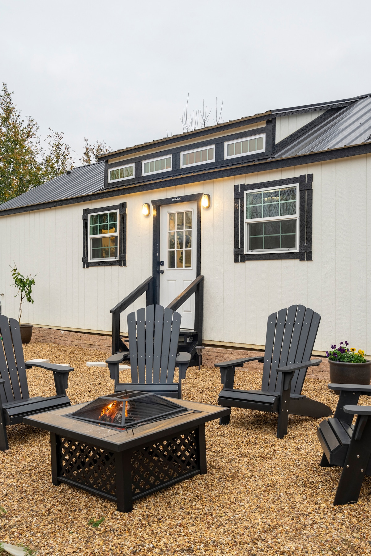An outdoor seating area features five black Adirondack chairs arranged around a fire pit on gravel. The house, with a white exterior and black trim, is visible in the background, highlighting large windows and a welcoming entrance.