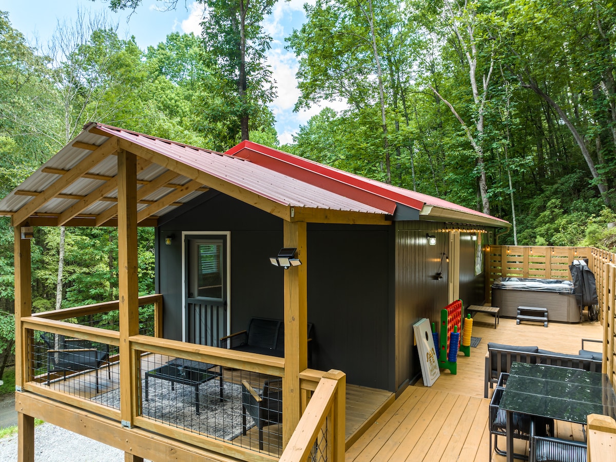 The cabin's exterior is set against a lush forest backdrop, featuring a covered porch with comfortable seating. A hot tub is located on the deck, while a dining area with a table and chairs is positioned nearby. Colorful outdoor games are visible.