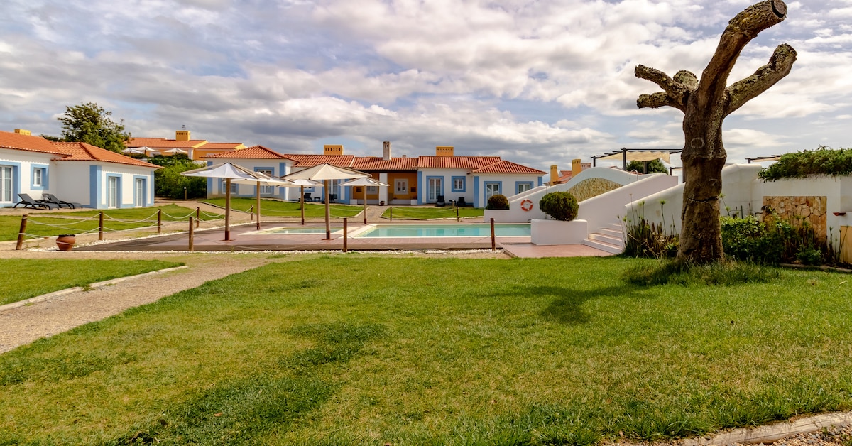 The outdoor pool is surrounded by well-maintained grass, with lounge chairs and umbrellas positioned nearby for relaxation. A charming tree with a unique shape stands prominently in the foreground, while the villas are visible in the background, featuring bright exteriors and inviting patios.