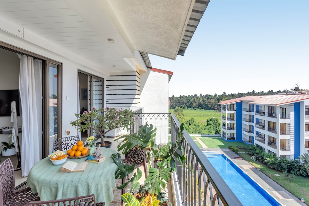 A balcony area features a round table covered with a light tablecloth, adorned with fruits and a plant. The view overlooks a shared swimming pool and lush greenery, creating a serene outdoor space. Elegant seating complements the area, inviting relaxation.
