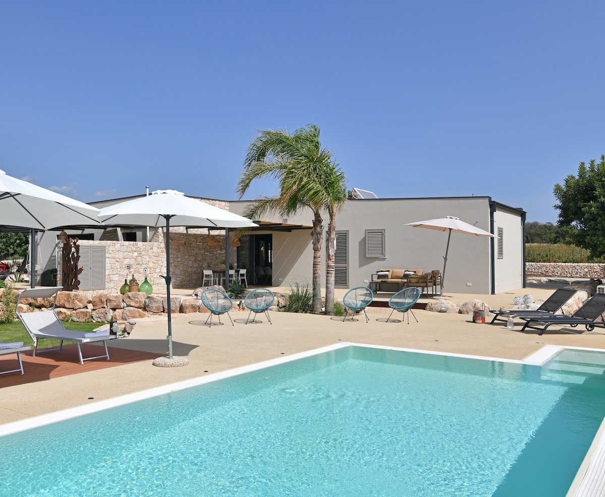 An outdoor area features a sparkling pool surrounded by sun loungers and umbrellas. Palm trees and greenery provide a refreshing atmosphere, while the modern structure offers a blend of indoor and outdoor spaces. A comfortable seating area is visible near the entrance.