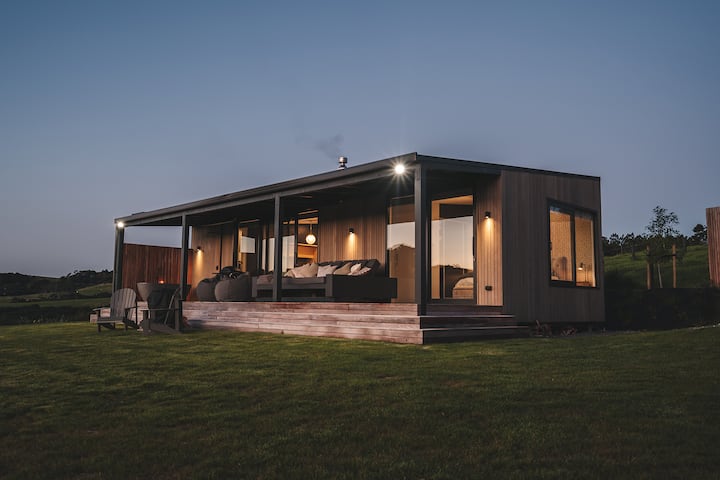 Modern cabin at dusk with a covered deck, cozy outdoor seating, warm interior lighting, and expansive glass windows