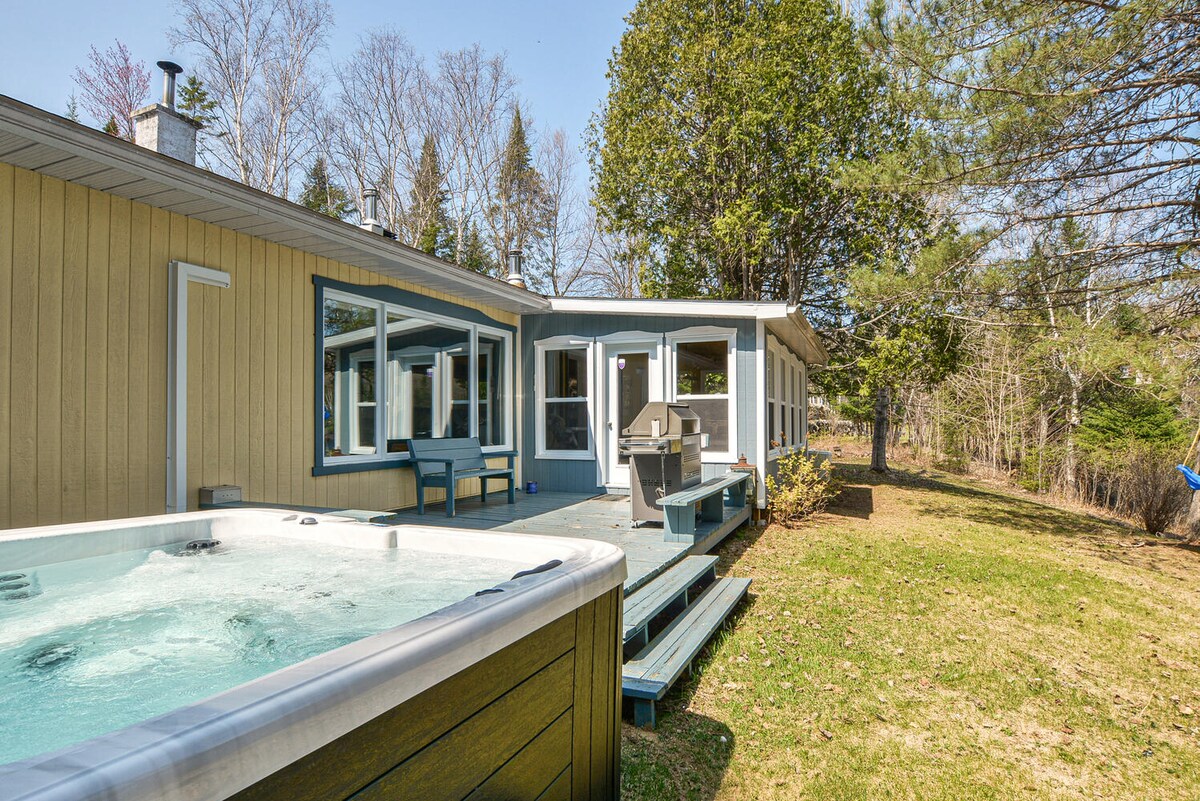 A private outdoor space features a hot tub positioned on a wooden deck. The area is surrounded by lush greenery and has a peaceful lawn. Large windows of the chalet provide natural light and views of the surrounding nature.
