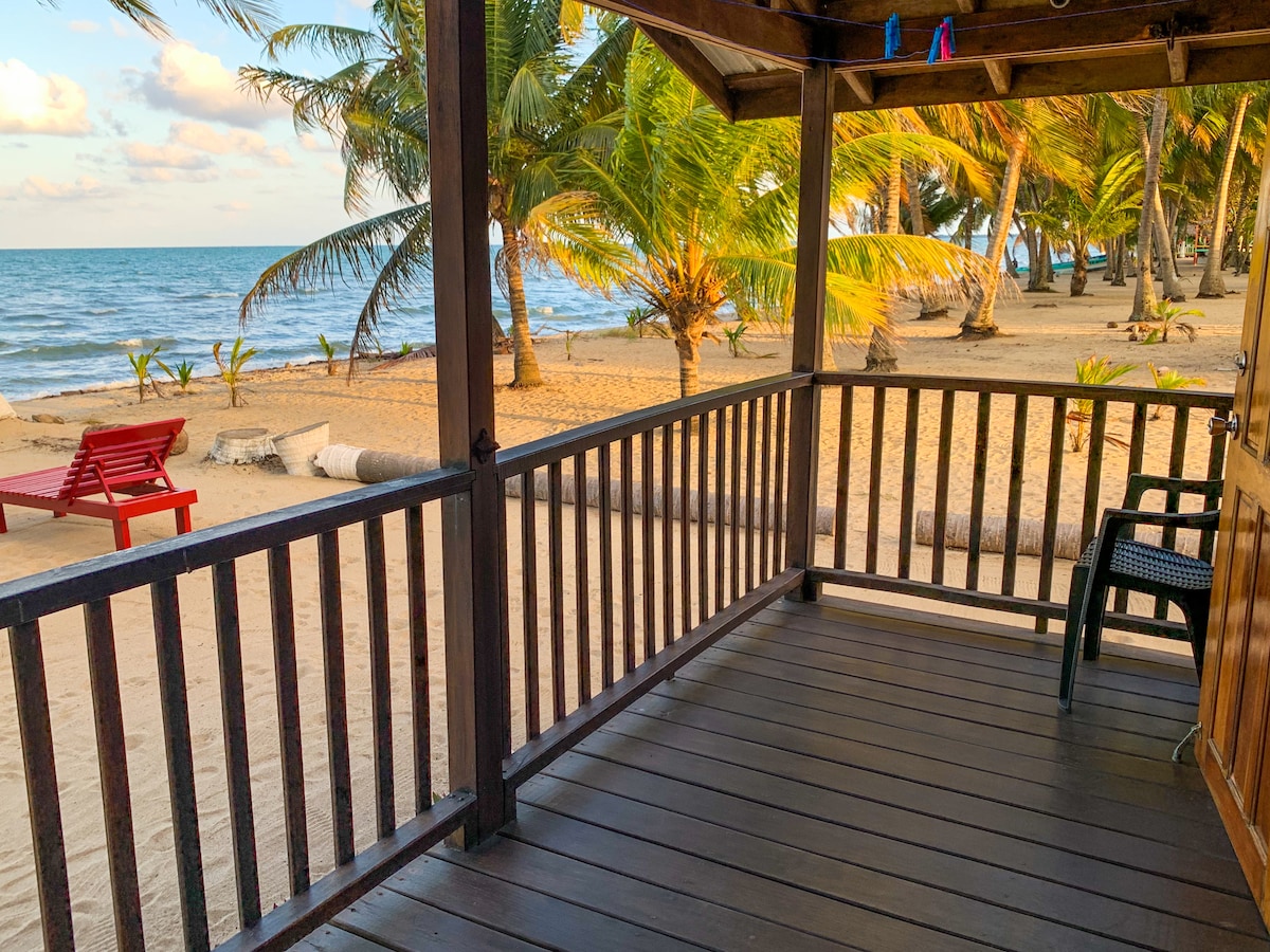 A wooden porch offers a view of the sandy beach and ocean, framed by palm trees. Red chairs are positioned on the beach, complementing the tranquil setting. The warm sun casts gentle shadows on the porch floor, creating an inviting outdoor space.