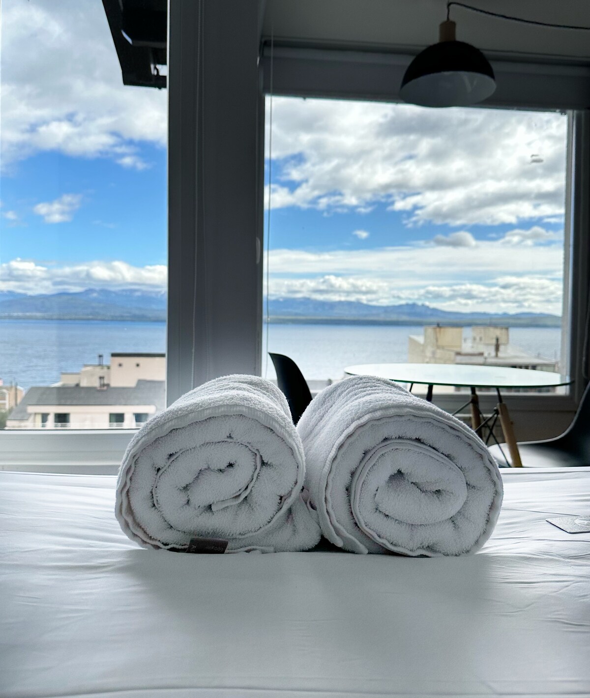 Two neatly rolled white towels are placed on a spacious bed. In the background, large windows offer a view of the expansive lake and mountains under a partly cloudy sky. A dining table and chairs are visible in the distance.