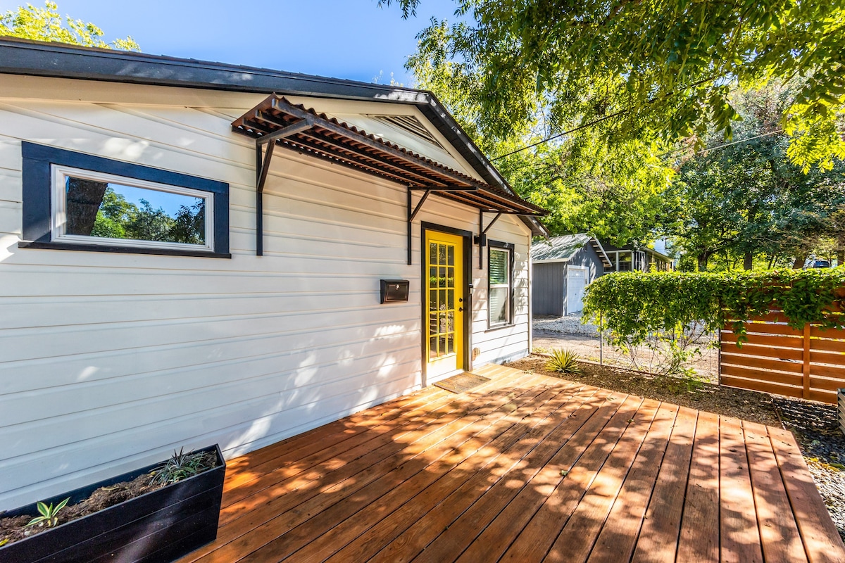 A welcoming exterior of a cottage features a wooden deck surrounded by greenery. A bright yellow door serves as a focal point, complemented by two windows on either side. The area is shaded by trees, creating a serene and inviting entrance.