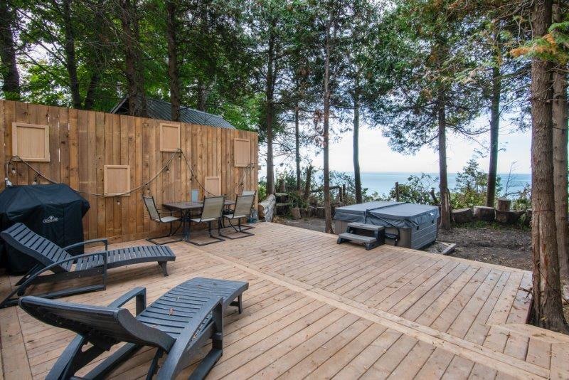 A spacious wooden deck is surrounded by trees, offering partial views of Lake Huron. Lounge chairs are positioned for relaxation, while a hot tub invites guests for a soak. A barbecue grill and outdoor dining table complement the inviting outdoor space.