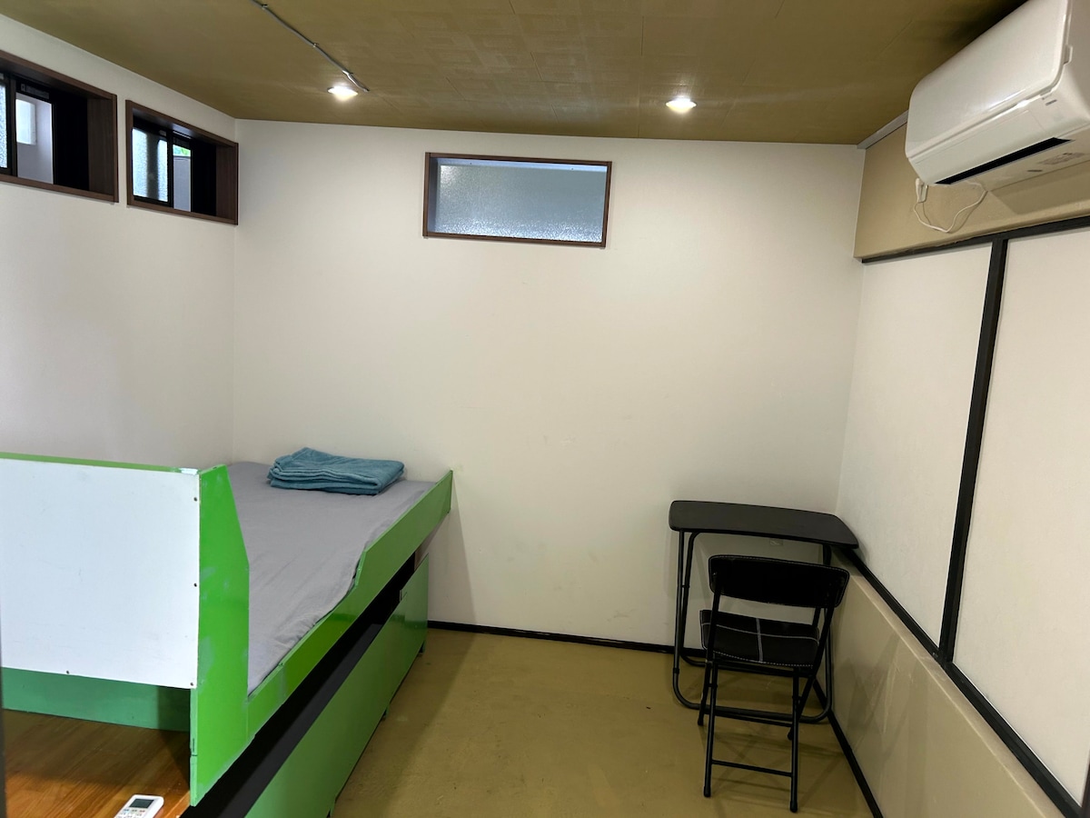 A simple sleeping area features a bed with a light grey blanket, positioned against a white wall. A small black chair and desk are located nearby. Natural light filters through a small window, enhancing the minimalistic design.