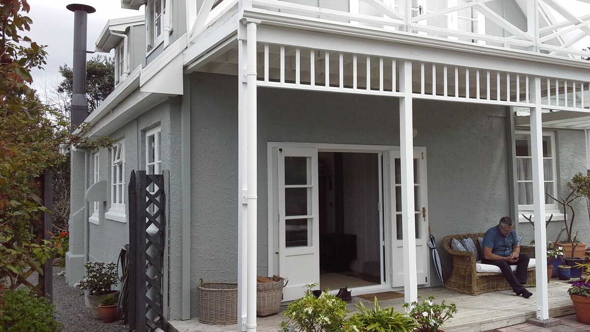 The exterior of a grey cottage is visible, featuring a spacious porch with white railings. A man is seated with a device, while potted plants and a decorative basket enhance the outdoor area. The structure includes large glass doors allowing natural light inside.