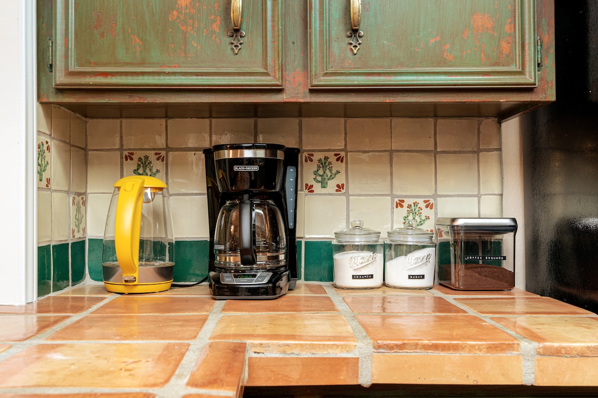 A cozy coffee bar is featured in the kitchen, showcasing a black coffee maker beside a yellow kettle. Glass containers hold sugar and ground coffee, positioned on a tiled countertop with a decorative backsplash. The rustic cabinet above adds a charming touch to the space.