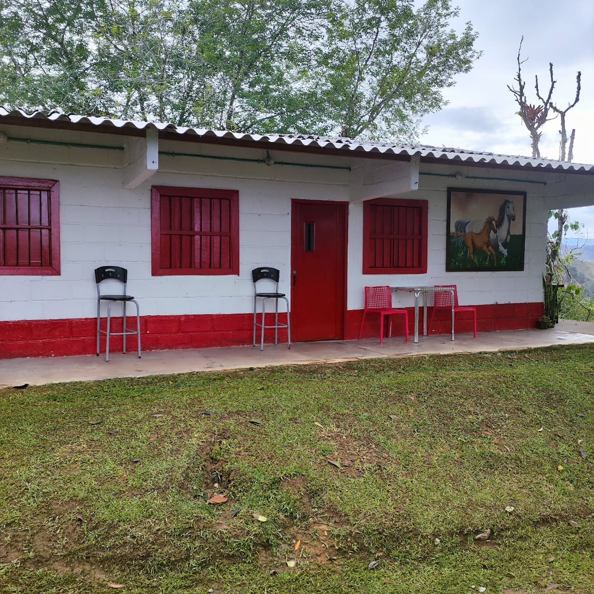 A charming countryside house with a red and white exterior is featured. Two tall chairs and a small table are arranged outside, offering a comfortable space to relax. Lush green grass surrounds the home, and trees provide a scenic backdrop.