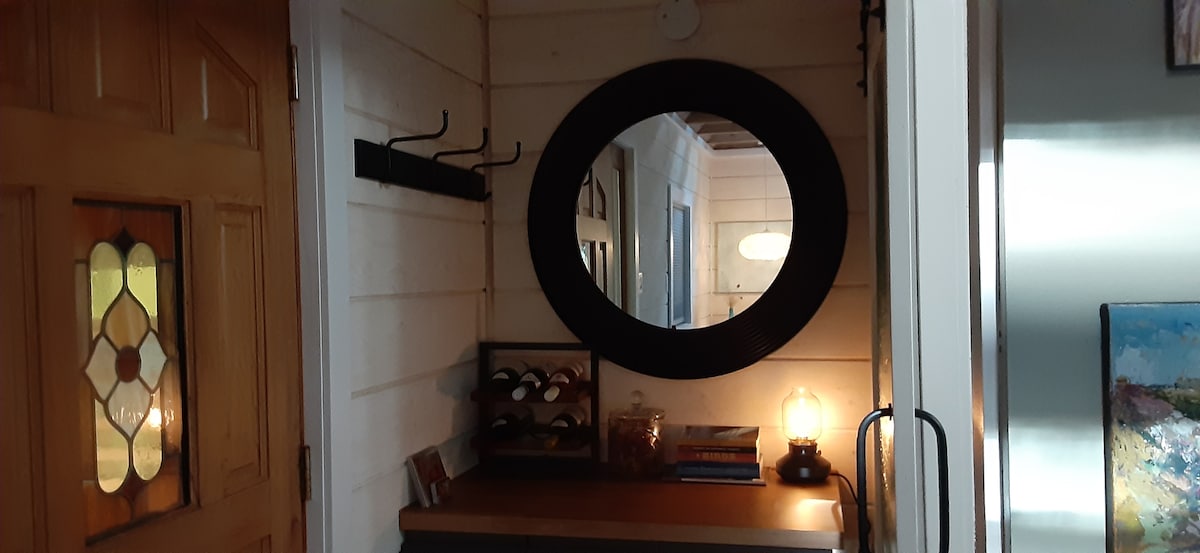 A welcoming entryway features a round mirror above a wooden console. A coat rack is mounted on the wall, and shelves hold a selection of wine bottles. Soft light is provided by a vintage-style lamp, enhancing the warm ambiance.