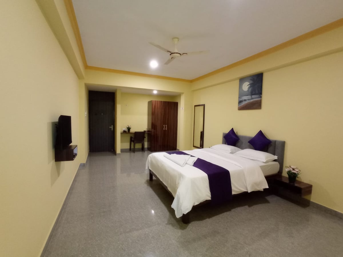 A spacious room is featured, displaying a queen-sized bed with purple and white linens. A television is mounted on the wall. A wooden table and two chairs are visible in the background, along with a mirror and decorative painting. Natural light fills the area.