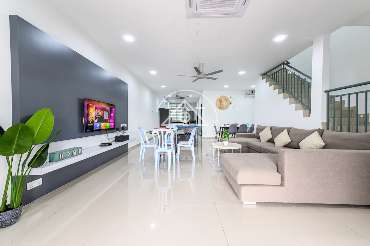 The living area features a modern design with sleek tile flooring and a large gray sectional sofa. A flat-screen TV is mounted on the wall opposite a dining area with white chairs. A ceiling fan is present, and a staircase leads to the upper level.
