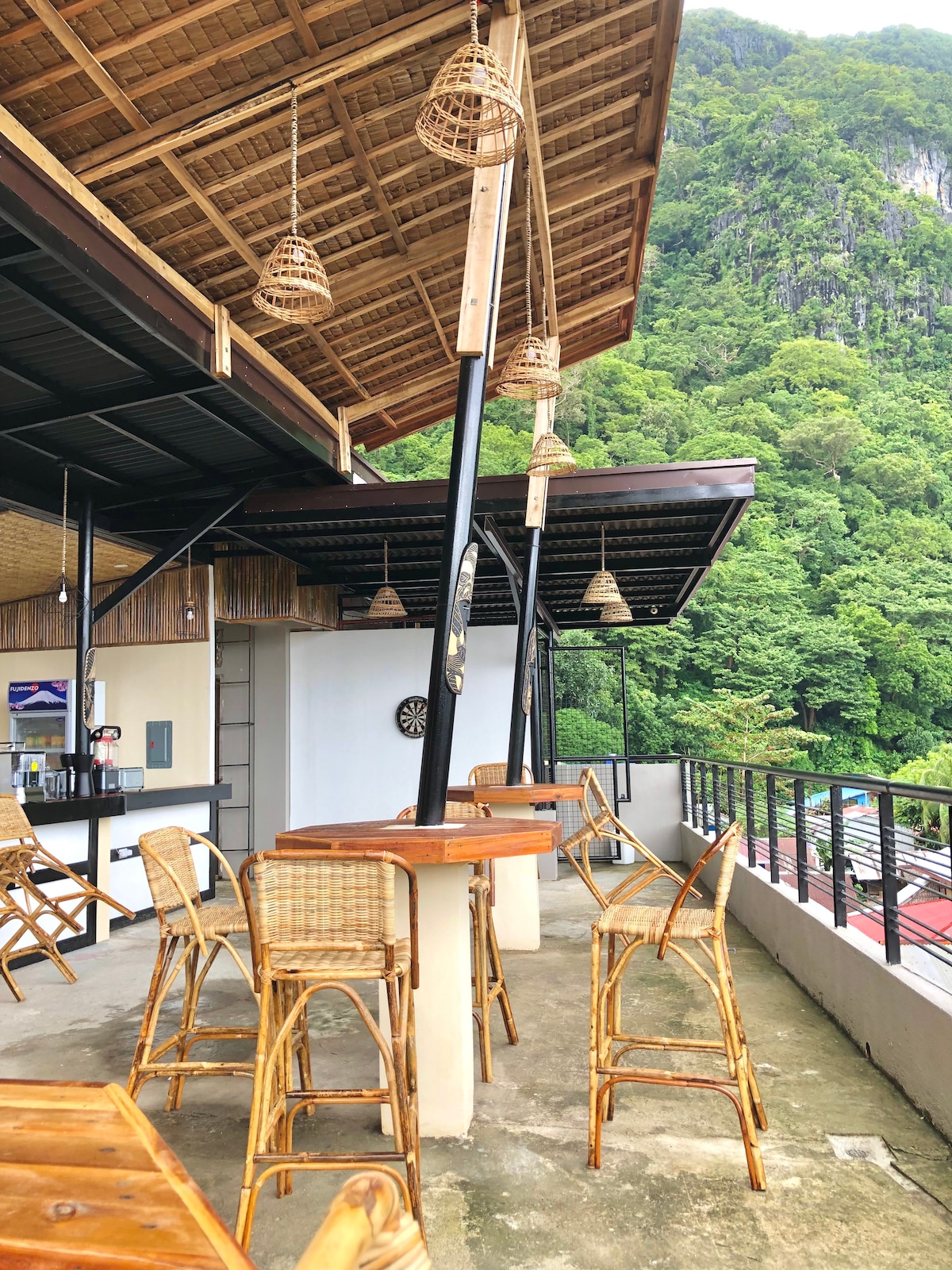An open-air rooftop area features wooden tables and tall woven chairs, creating a casual dining setting. Natural light flows into the space, highlighting the bamboo accents and surrounding greenery. The backdrop includes a lush hillside, adding a serene touch to the environment.