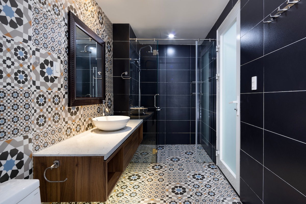 The bathroom features a modern design with patterned tiles and a spacious layout. A large mirror is mounted above the vanity, which includes a vessel sink. A glass-enclosed shower is visible, complemented by dark cabinetry and a neutral color palette.