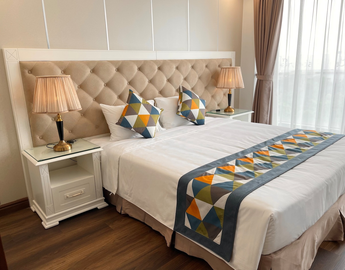 A comfortable bedroom features a spacious bed with a soft headboard and decorative pillows. Two bedside tables with lamps are positioned on either side. Light filters through sheer curtains, illuminating the neutral-colored walls and modern décor.