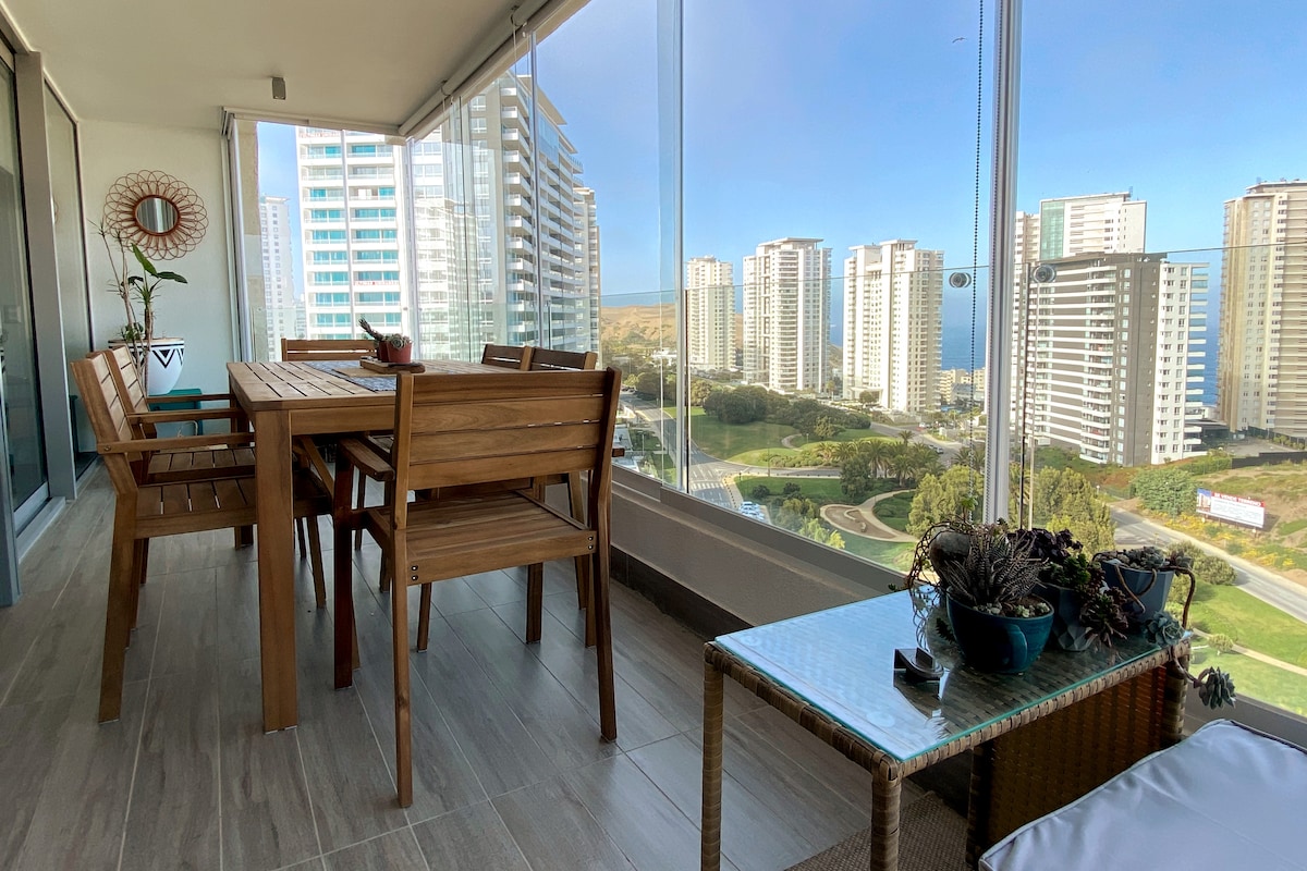 A spacious terrace is featured, overlooking a park and cityscape. A wooden dining table with matching chairs is visible alongside a wicker coffee table adorned with potted plants. Floor-to-ceiling glass windows provide a panoramic view, allowing natural light to fill the area.