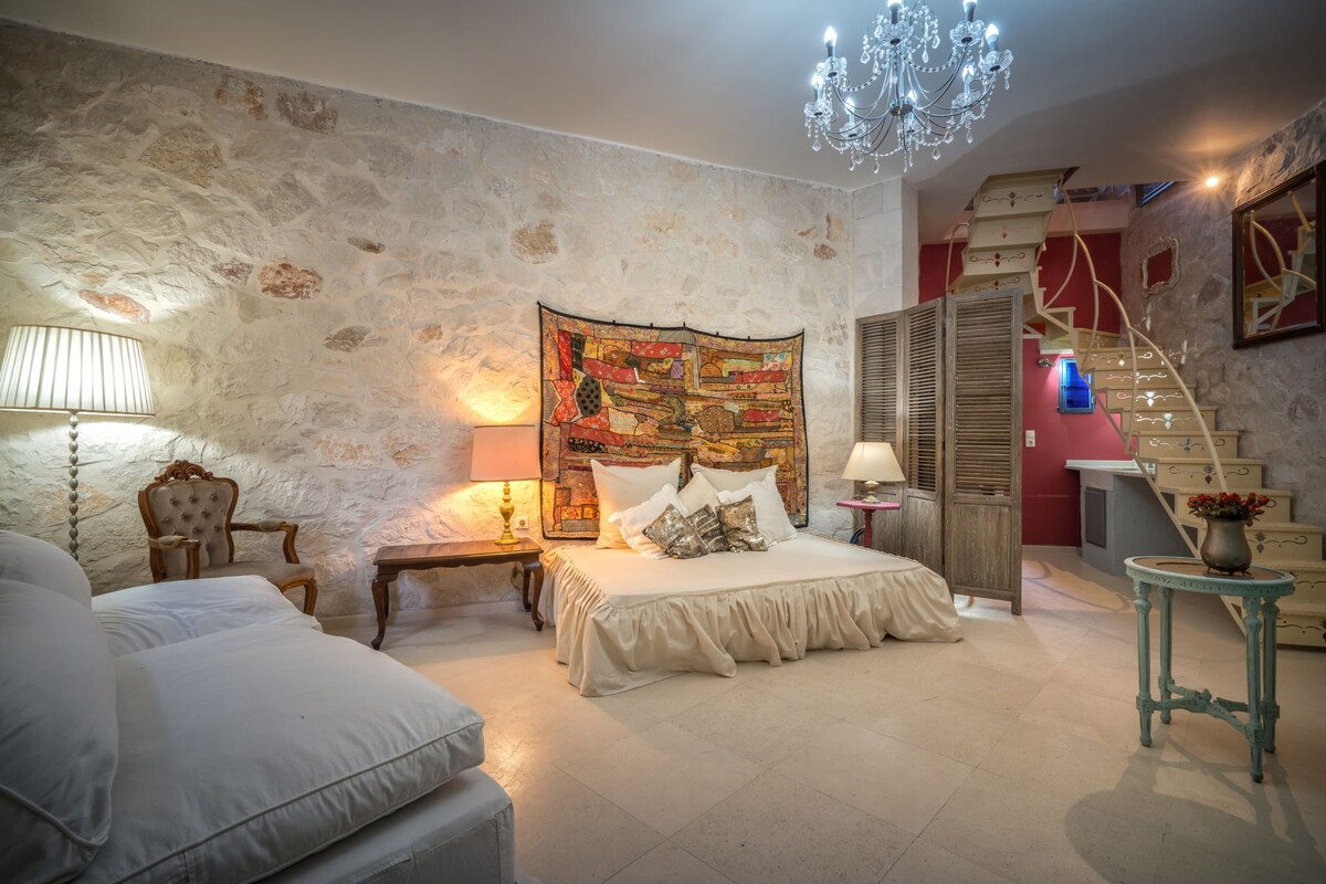 The spacious living area features a large double bed with soft linens and a decorative wall tapestry. A vintage armchair and small table are positioned nearby. A staircase leads to the upper floor, while natural light brightens the room through a modest window, complemented by elegant lighting.