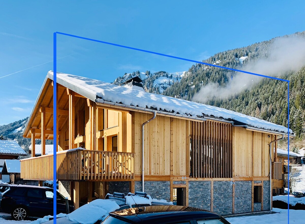The chalet features a wooden exterior with a sloping roof, surrounded by mountains. A spacious outdoor area offers a wooden deck with seating. Large windows provide views of the natural landscape, while the gray stone base complements the wooden structure.