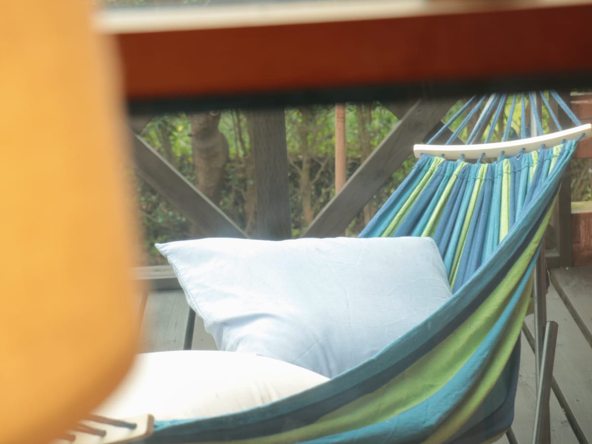 A colorful hammock rests on a balcony, bordered by wooden railings and surrounded by greenery. A light blue pillow adds comfort, while soft natural light filters in through the nearby window.