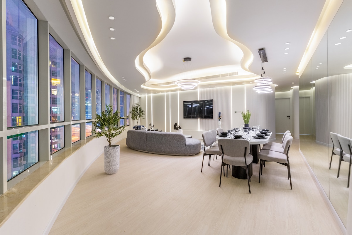 The open plan living and dining area features a spacious layout with large windows, providing a view of the city. A modern sofa and lounge chair are complemented by a marble dining table that seats eight. Natural elements, including a potted plant, add a touch of greenery.