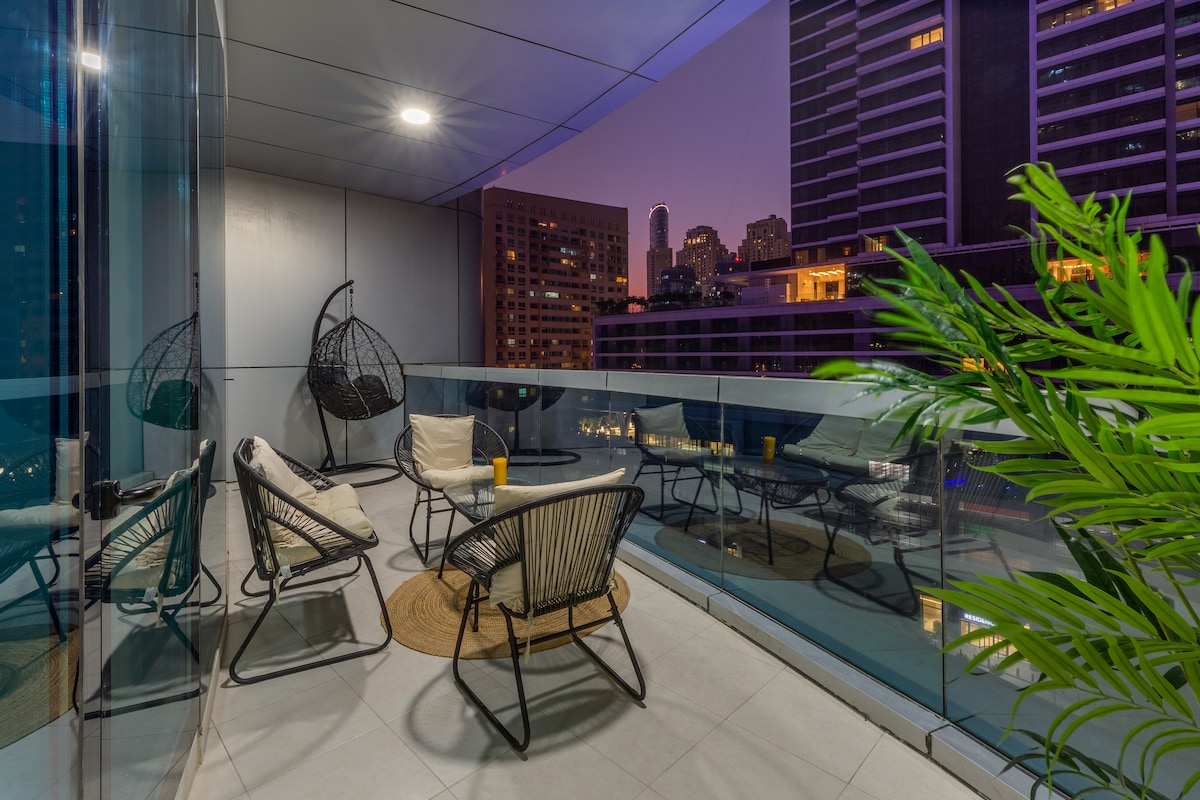 A furnished balcony features a cozy seating arrangement with three lounge chairs and a round table. A circular hanging chair adds a unique touch. The space is illuminated by evening lights, with a view of the city skyline in the background.