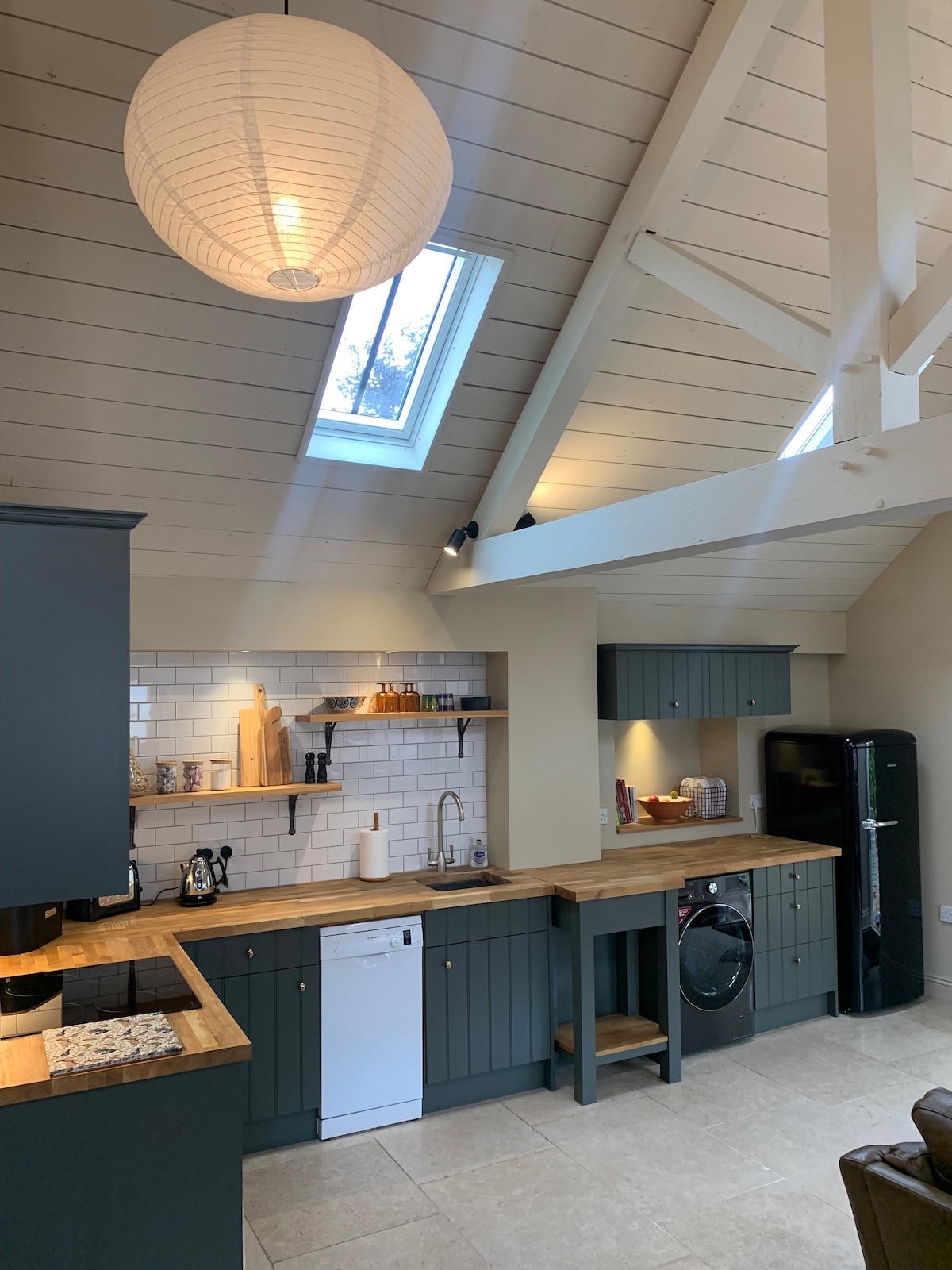 A modern kitchen area is presented with wood countertops and dark cabinetry. Skylights allow natural light to fill the space, highlighting the contemporary appliances including a dishwasher and refrigerator. The open design features a charming blend of functionality and style.
