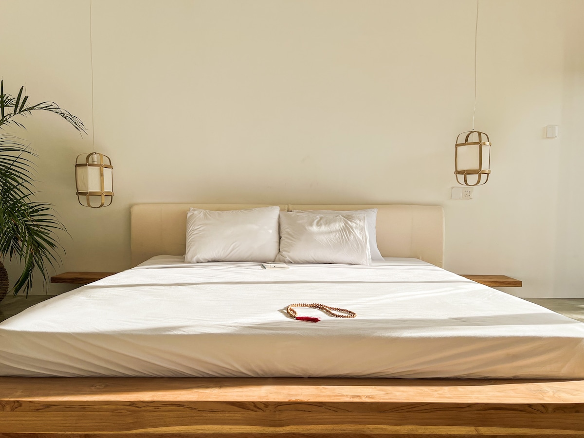 A spacious bed with a light-colored headboard is centered against a neutral wall. Two softly glowing pendant lights are positioned above the nightstands. A decorative item rests on the bed, and a potted plant adds a touch of greenery to the corner.