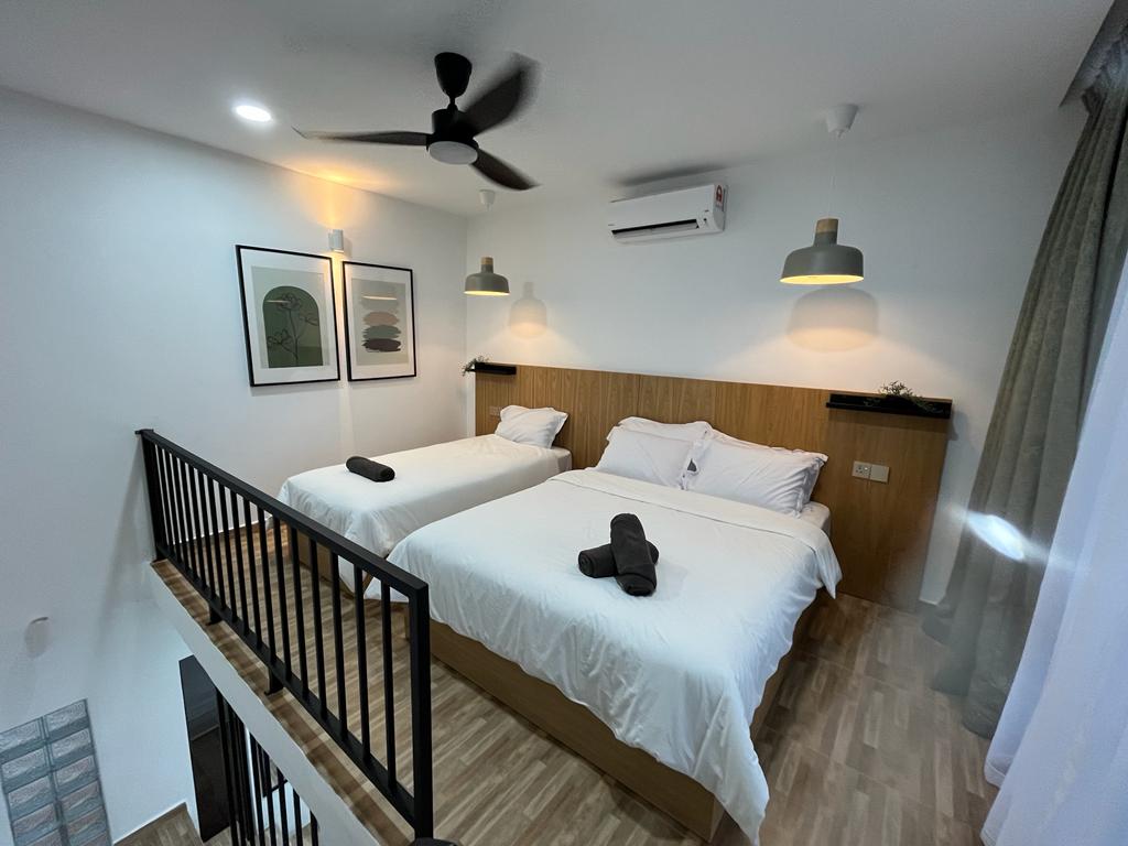 Two spacious beds are positioned in a bright, airy room with modern decor. Soft linens and dark towels are neatly arranged on each bed. Large windows provide natural light, while a ceiling fan circulates air. Simple artwork and wall lighting add a contemporary touch.