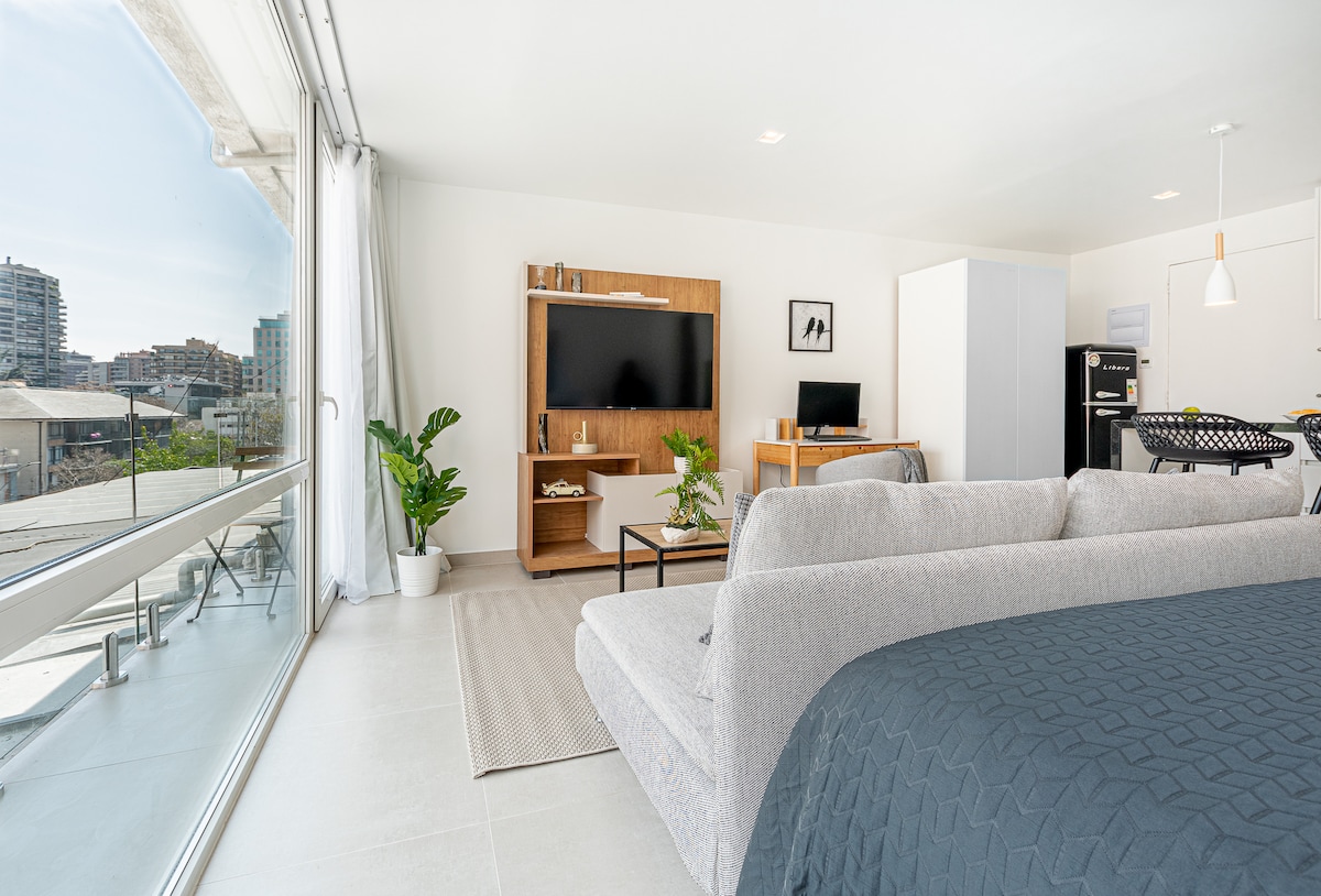An open living space features a comfortable sofa and a large television mounted on a wooden media console. Natural light enters through expansive glass doors, while a small desk with a monitor is visible. A decorative plant adds a touch of greenery to the scene.