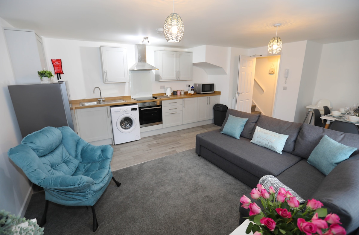 The modern kitchen area features light cabinetry and a wooden countertop. Appliances include a washing machine, microwave, and oven. A comfortable grey sectional sofa with teal pillows is positioned in the inviting lounge area, while a bouquet of roses adds a touch of color to the scene.