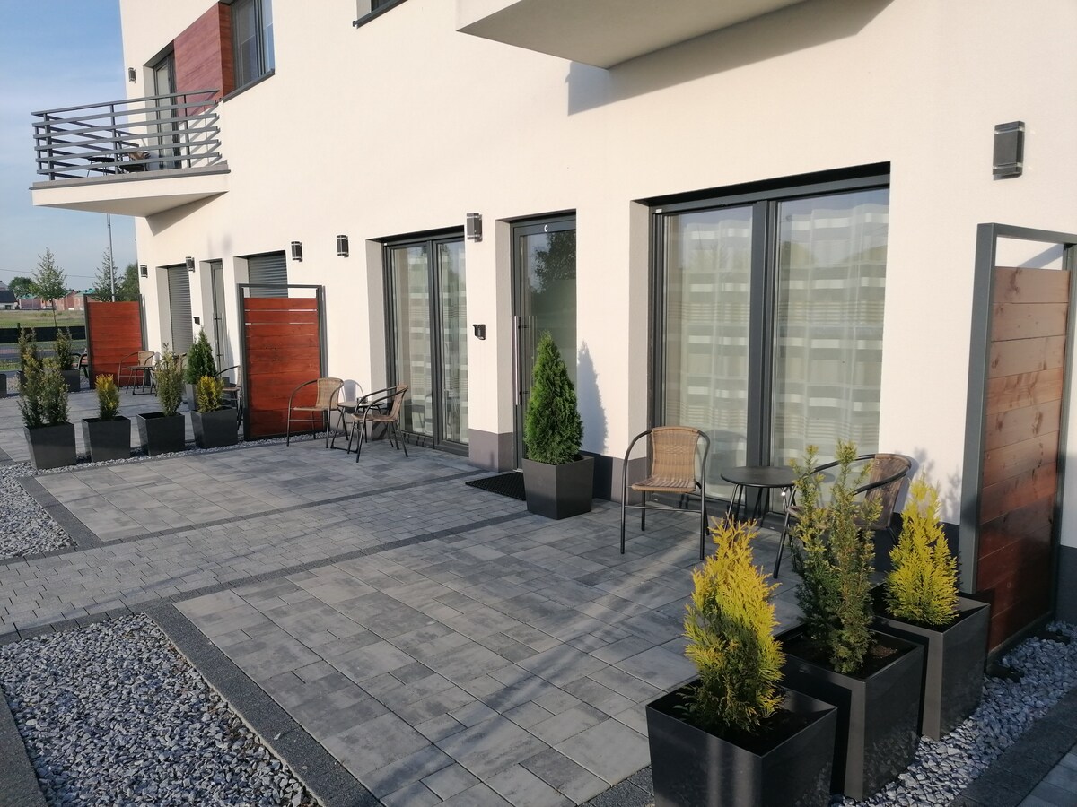 An outdoor area is featured with a paved surface and several planters containing greenery. Seating arrangements include two chairs positioned near the building's entrance. Sliding glass doors offer access to indoor spaces, framed by contemporary architecture.