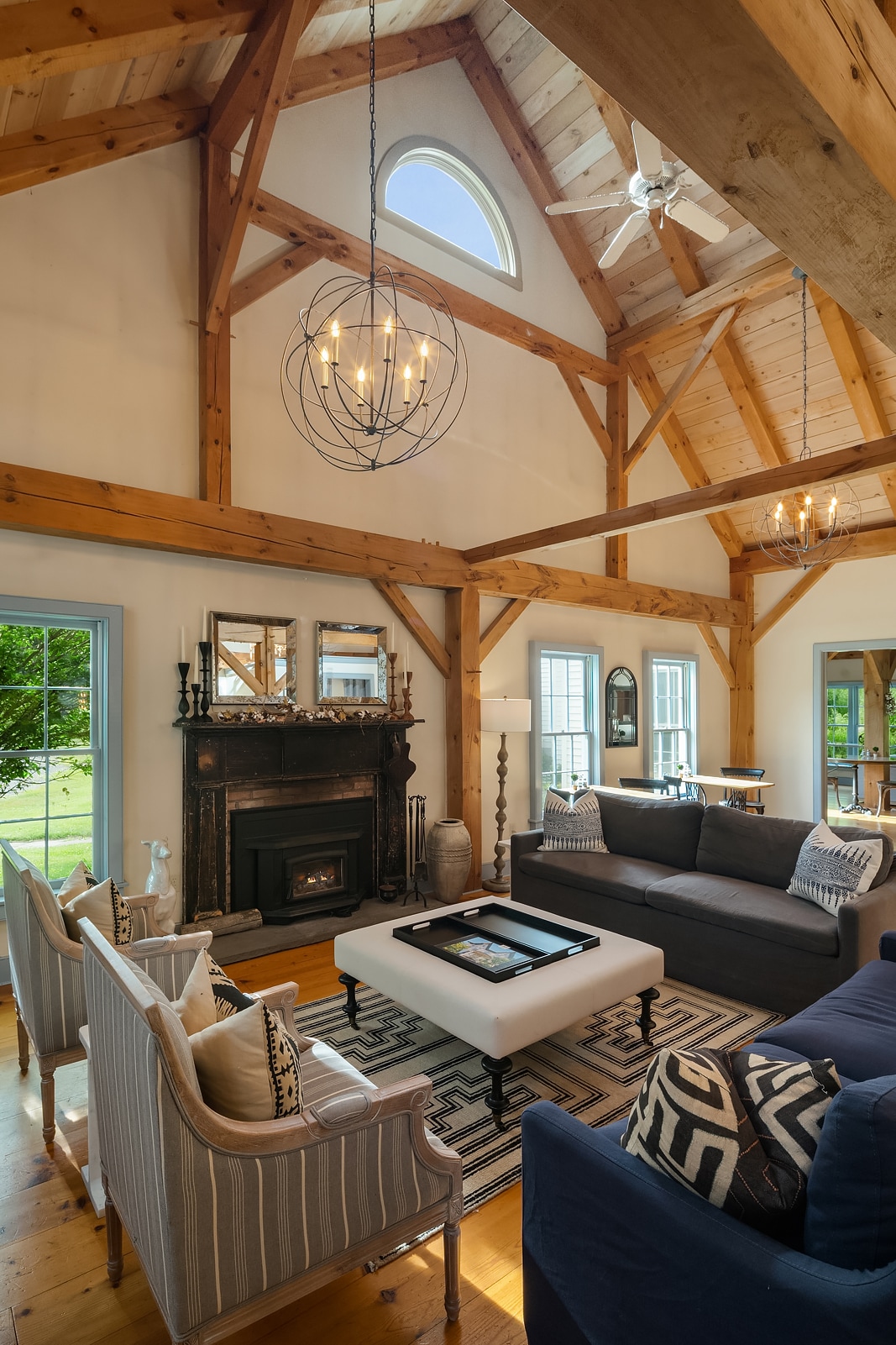 The living area showcases high, vaulted ceilings with exposed wooden beams and large windows allowing natural light to fill the space. Comfortable seating includes a dark sofa and two patterned armchairs, arranged around a central coffee table. A decorative fireplace adds to the room’s charm.