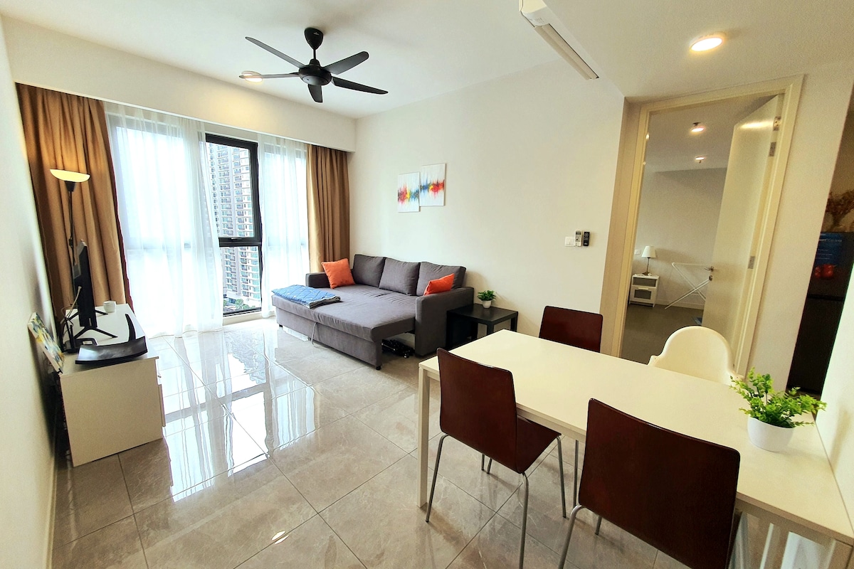 A contemporary living area features a spacious sectional sofa and a small dining table surrounded by four chairs. Large windows allow natural light to fill the room, and light-colored curtains frame the view. A flat-screen TV is positioned on a cabinet across from the seating.