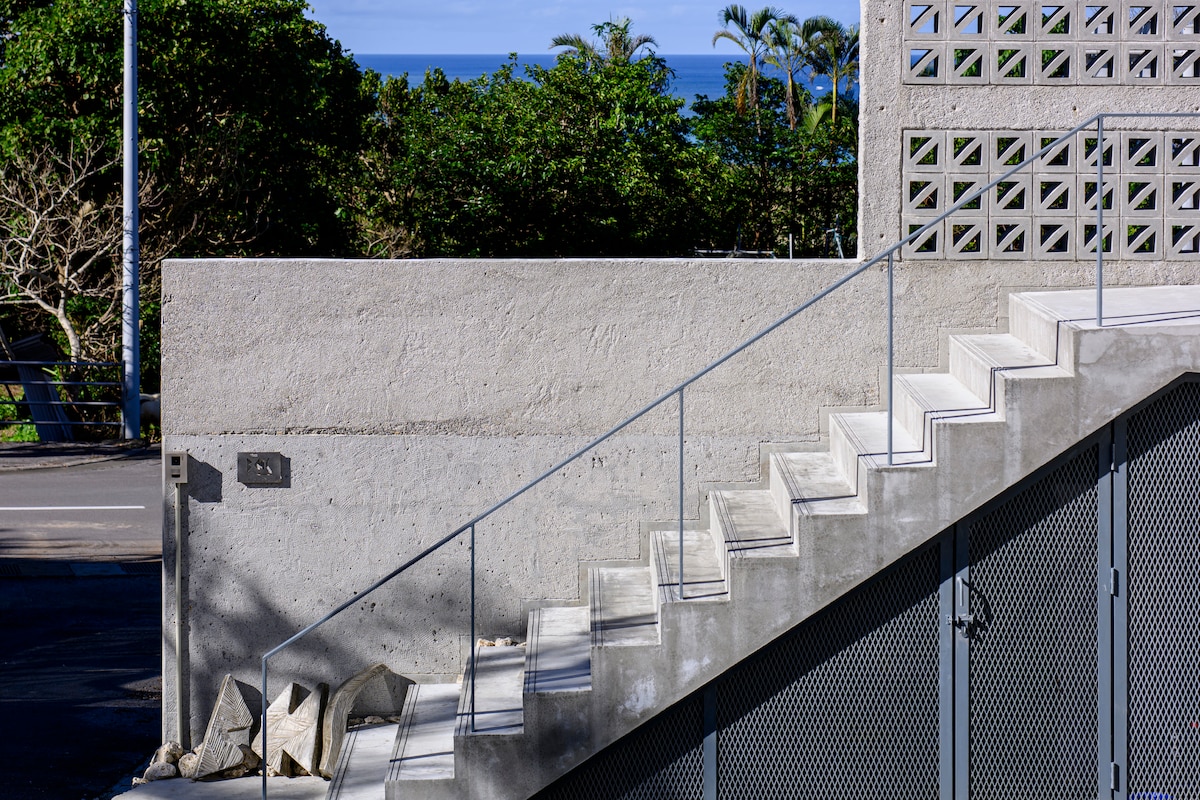 A concrete staircase leads towards a textured wall, surrounded by greenery with the ocean visible in the background. The sturdy metal railing complements the minimalist design, creating a functional yet elegant pathway. Sunlight casts gentle shadows, accentuating the architectural elements.