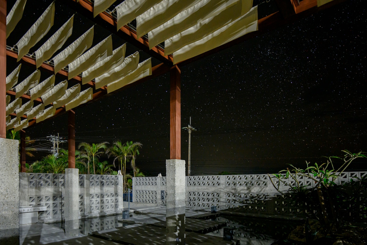 An outdoor terrace is depicted under a star-filled sky, showcasing wooden beams and fabric canopies that provide shelter. The area features textured concrete walls and lush greenery, creating a serene and open atmosphere. The surrounding darkness enhances the visibility of the twinkling stars above.