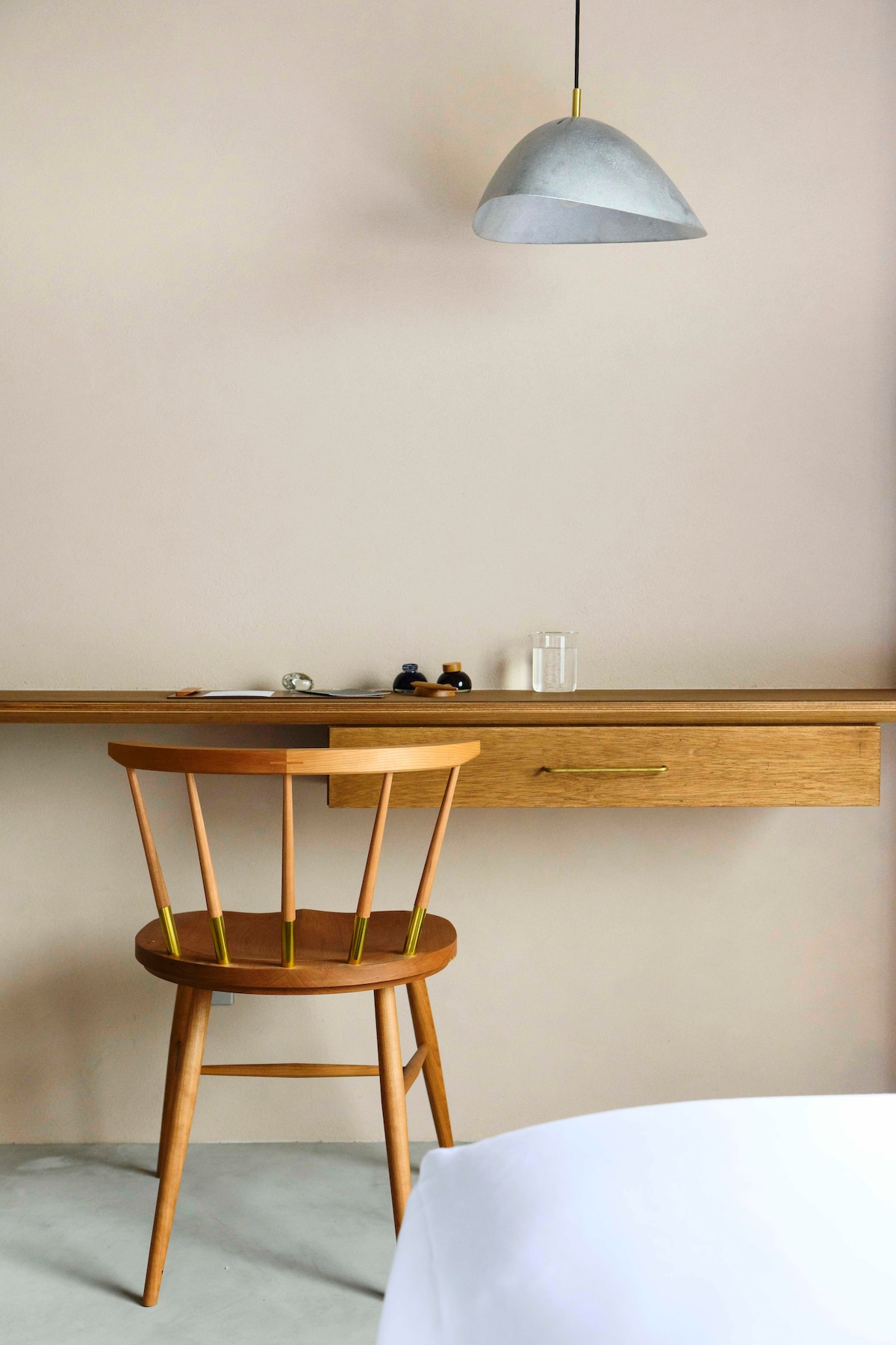 A minimalistic workspace is presented with a wooden desk and a matching chair, positioned against a soft, pale wall. A subtle gray pendant light hangs above, while a few decorative items sit on the desk, enhancing the serene environment.