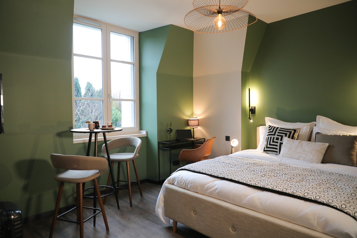 A well-appointed bedroom features a spacious bed with neutral bedding. A cozy armchair and small table are positioned near a window that offers a view of the outdoors. Soft, green walls complement the natural light and add a serene touch to the room.