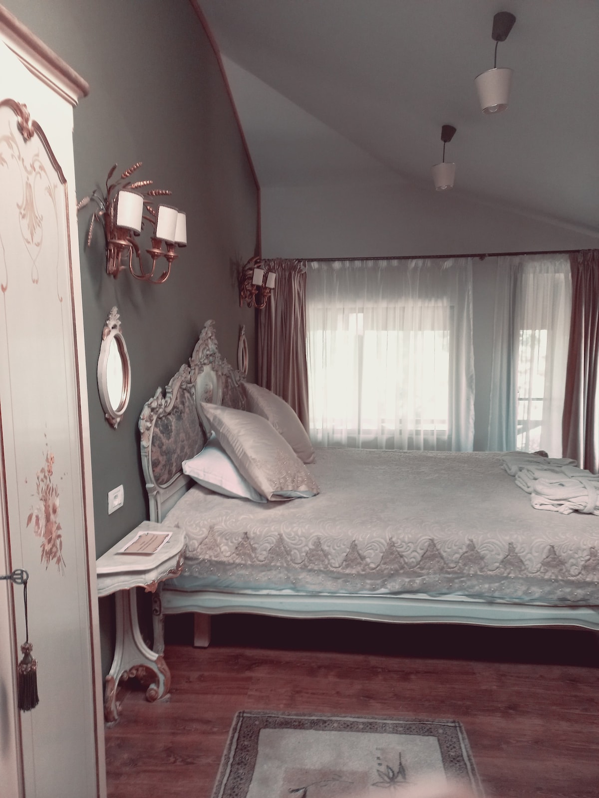 A serene bedroom features a spacious bed with light bedding and decorative pillows. Soft natural light enters through large windows, accentuated by flowing curtains. An ornate wardrobe and bedside table complement the elegant decor, while wall sconces add gentle illumination.
