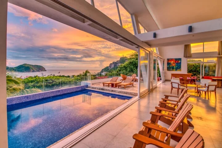 An open living area with large glass doors features a pool with a reflective surface, set against a backdrop of a vibrant sunset over the ocean. Stylish wooden deck chairs are positioned for viewing the scenic landscape, creating an inviting space for relaxation.