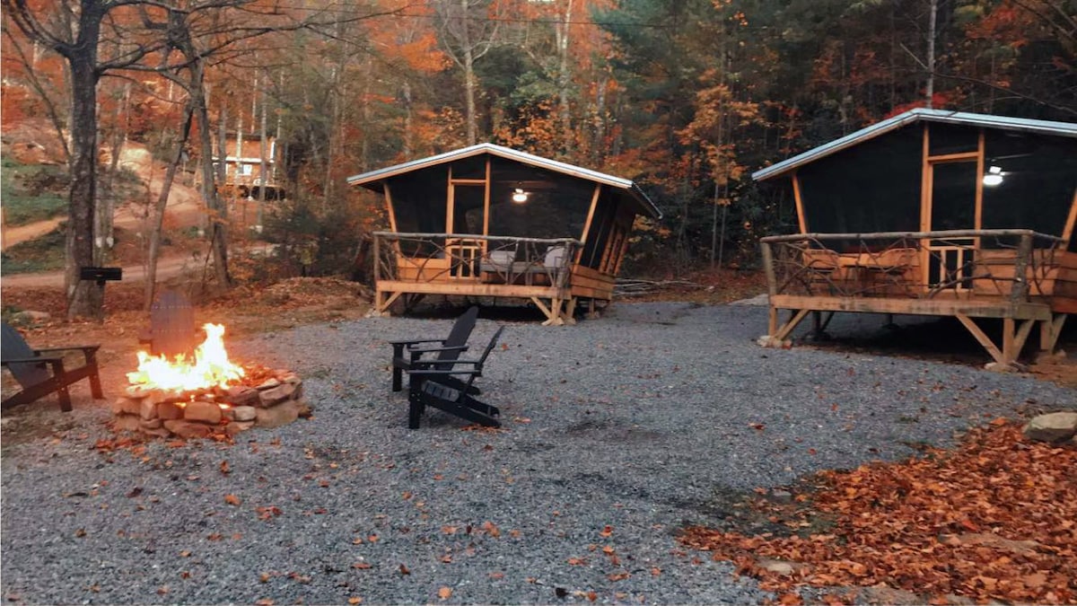 Two glamping cabins are positioned near a gravel area, surrounded by autumn foliage. A central fire pit, surrounded by stone, is filled with flames, providing visual warmth. Each cabin features an outdoor deck with chairs and a table, offering an inviting space for relaxation.