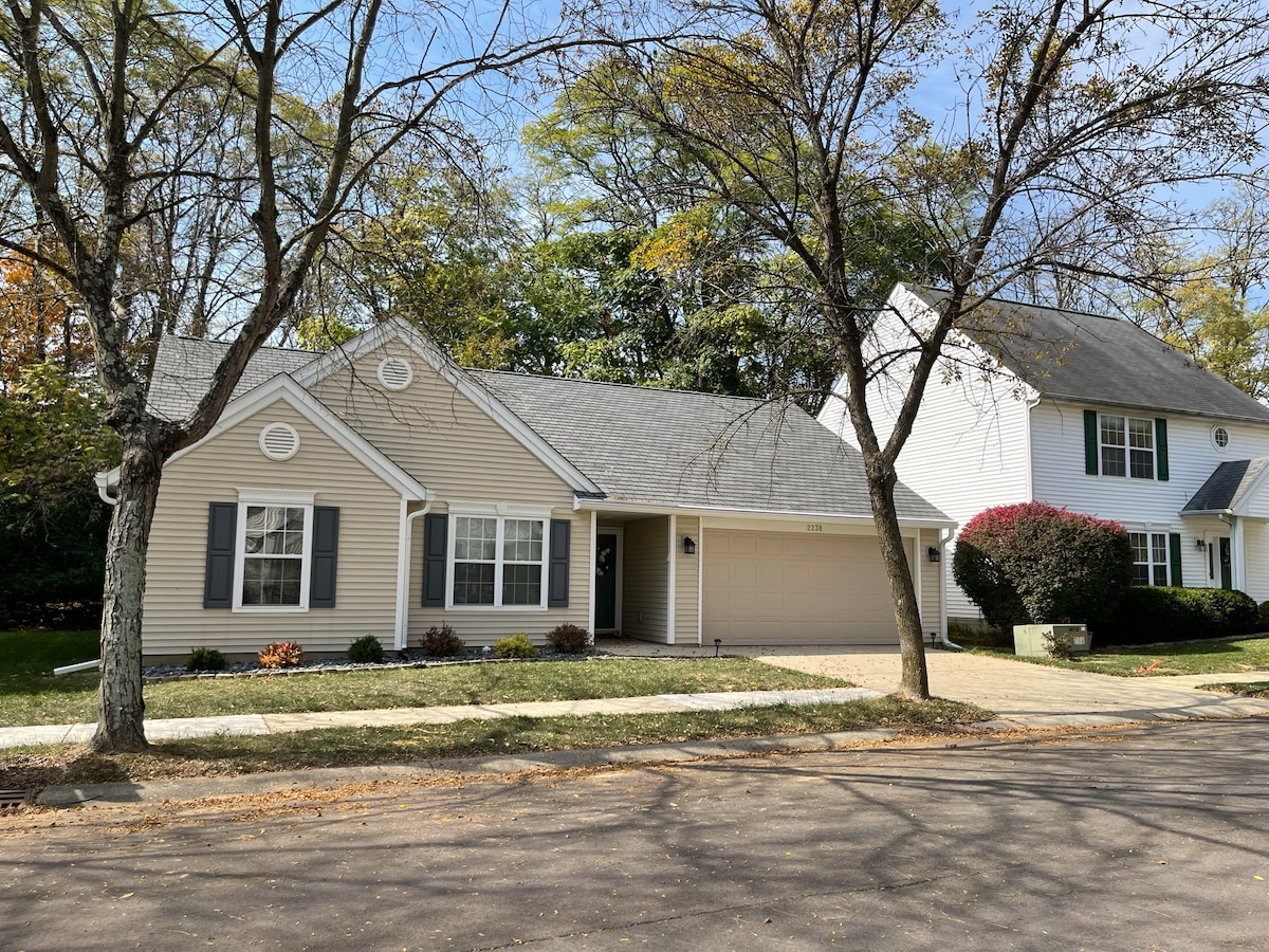 A single-family home is featured, showcasing a light exterior with dark shutters. The front yard is well-maintained, and a paved driveway leads to the attached garage. Surrounding trees and landscaped greenery enhance the neighborhood's peaceful atmosphere.
