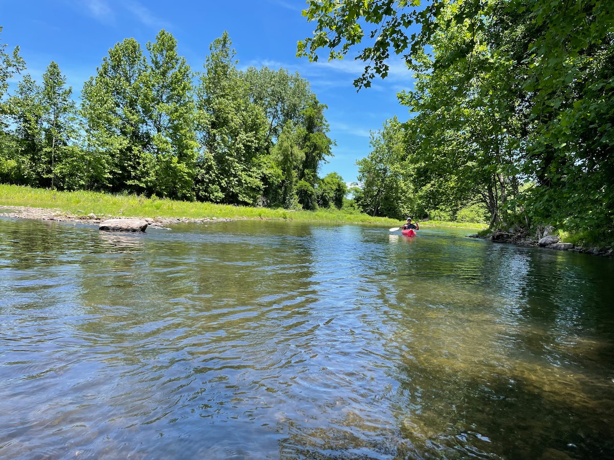 A tranquil river scene is displayed, with clear water reflecting the blue sky and lush greenery on both banks. Verdant trees provide shade along the water's edge, while a kayaker is seen paddling peacefully in the distance.