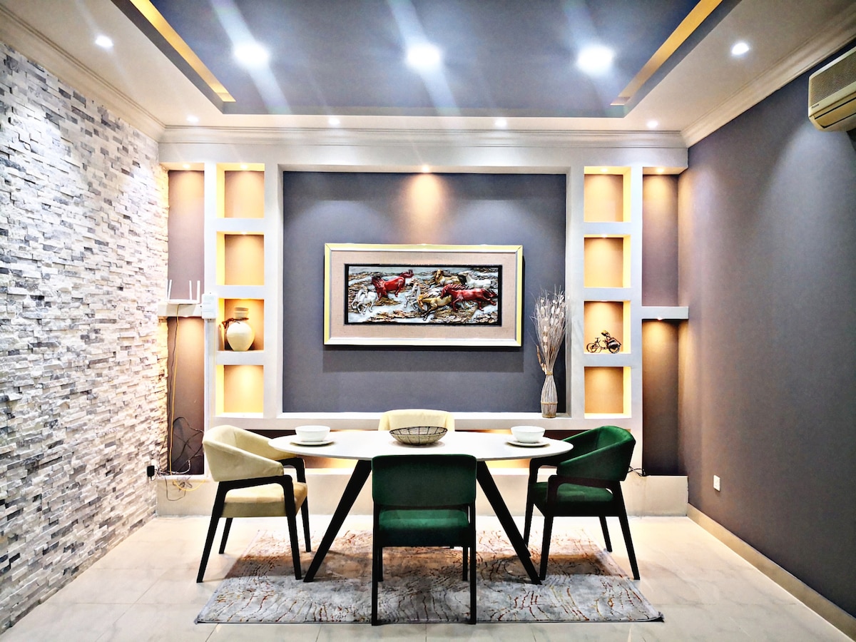 A dining area features a large table surrounded by four chairs, two in green and two in neutral tones. A textured stone wall adds contrast, while recessed lighting highlights a central artwork. The space is designed for a modern, inviting atmosphere.