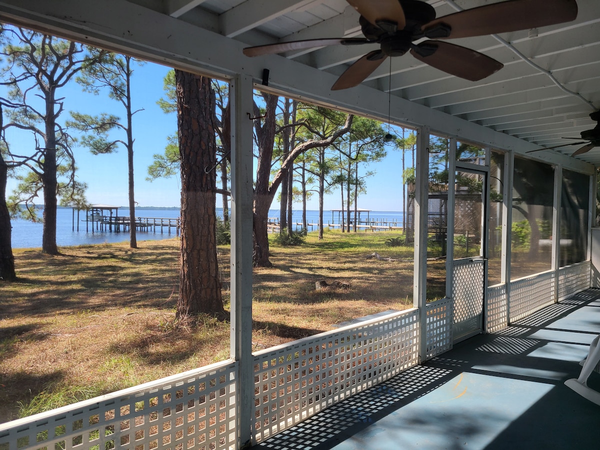 A screened-in porch features a view of a calm bay, framed by tall pine trees. Natural light floods the space, highlighting the wood ceiling and ceiling fans. A wooden pier extends into the water, inviting relaxation and enjoyment of the serene surroundings.