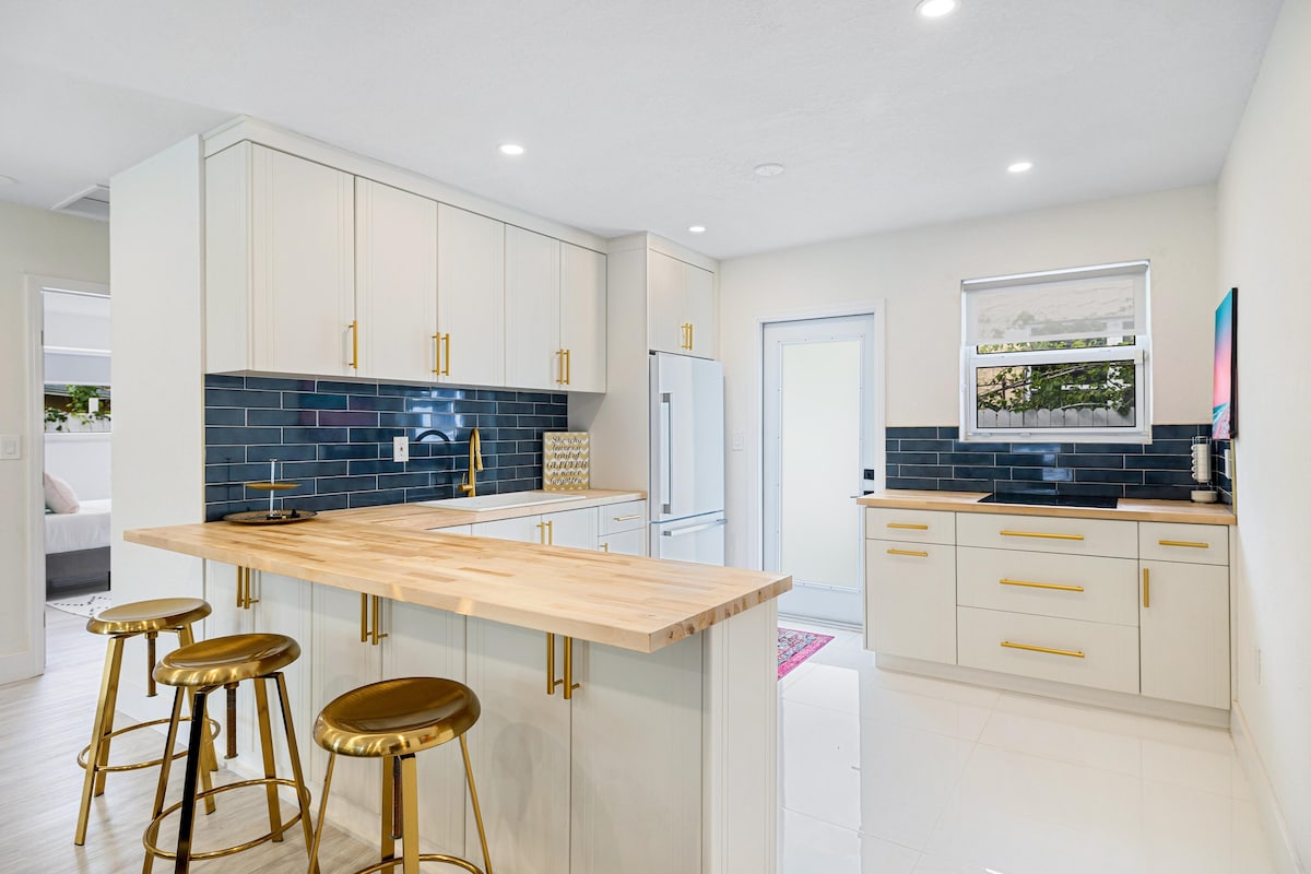 A modern kitchen features light-colored cabinetry with gold hardware, complemented by a sleek blue backsplash. A wooden island with three metal stools offers a casual dining option. Natural light brightens the space, enhancing the contemporary appliances and polished floors.