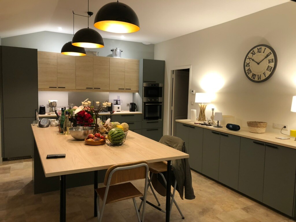 A modern kitchen features a central island with a light wooden countertop, surrounded by chairs. Dark cabinets complement the light walls. Various kitchen appliances and a decorative arrangement of fruits are visible. Warm pendant lights illuminate the space, creating a bright and functional atmosphere.