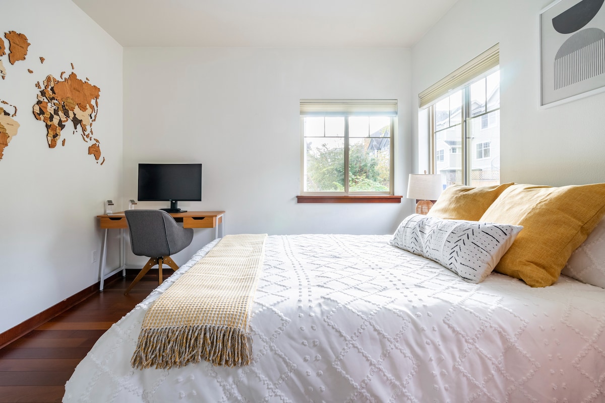 A bright bedroom features a spacious bed adorned with neutral-colored bedding and a decorative throw. A simple desk with a computer is positioned nearby, complemented by a comfortable chair. Natural light streams through a window, highlighting a world map art piece on the wall.