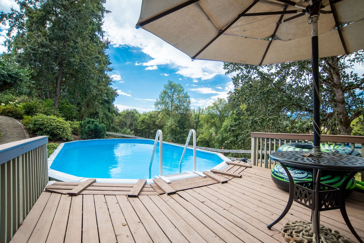 A private pool is surrounded by wooden decking and lush greenery. An umbrella offers shade over a table and chairs, creating a space for relaxation. Steps lead into the water, which reflects the clear blue sky and surrounding trees.